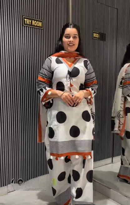 A woman modeling a white three-piece salwar kameez suit with black polka dots, gray stripes, and orange accents, standing in front of a gray slatted wall.