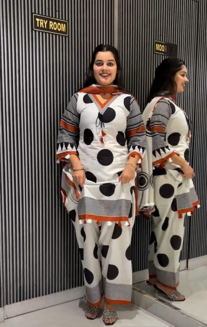 A woman modeling a white three-piece salwar kameez suit with black polka dots and orange accents, standing in front of a gray striped wall with a mirror showing the back view.