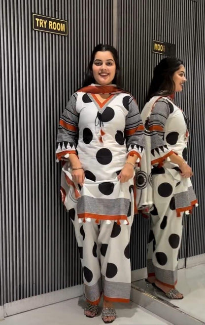 A woman modeling a white three-piece salwar kameez suit with black polka dots and orange accents, standing in front of a gray striped wall with a mirror showing the back view.