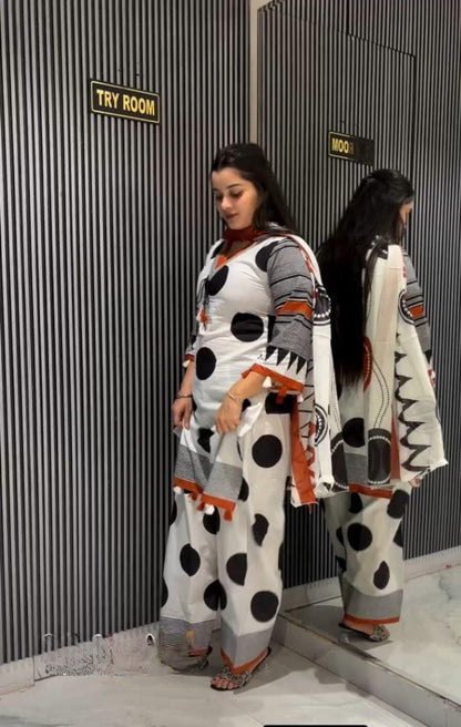 A woman stands in a fitting room wearing a white kurta and wide-leg pants set with black polka dots and gray patterned sleeves and hem, alongside her reflection in a mirror.