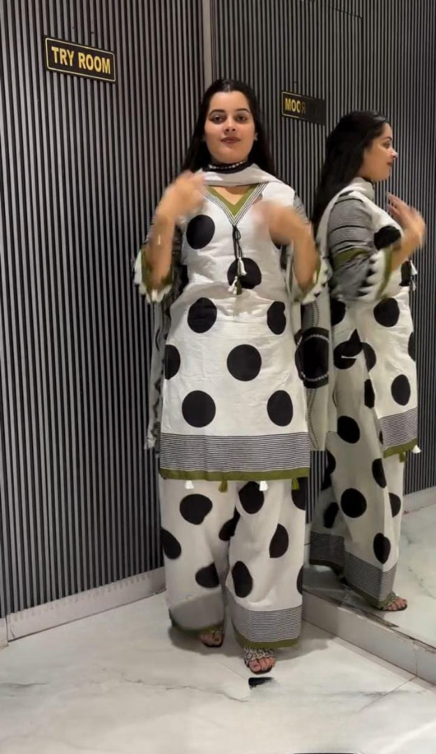 A woman modeling a white three-piece salwar kameez suit with large black polka dots and gray striped borders, standing in front of a mirror that shows the back view of the outfit.