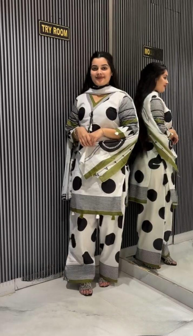 A woman modeling a white three-piece salwar kameez suit with black polka dots and olive green stripes, standing in front of a mirror that reflects her side profile. The background is a gray corrugated metal wall with a 'TRY ROOM' sign.