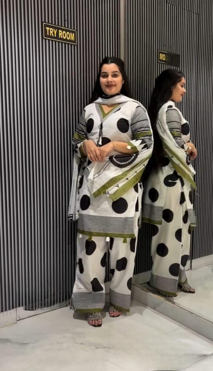 A woman modeling a white three-piece salwar kameez suit with black polka dots and olive green stripes, standing in front of a mirror that reflects her side profile. The background is a gray corrugated metal wall with a 'TRY ROOM' sign.