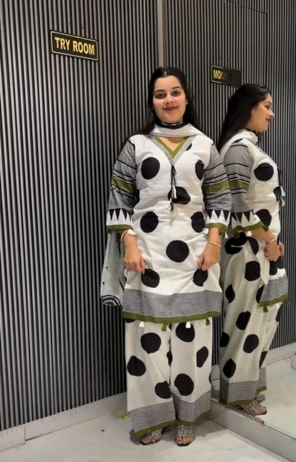 A woman modeling a white three-piece traditional outfit with large black polka dots, gray patterned sleeves and hem, and olive green accents, standing in front of a gray corrugated wall with a mirror showing the back view.