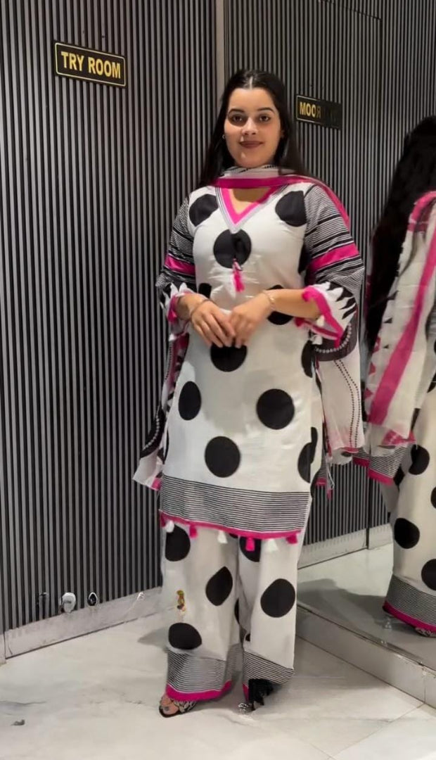 A woman modeling a white three-piece kurta set with large black polka dots and bright pink accents, standing in front of a gray striped wall with a mirror.