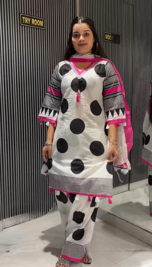 A woman modeling a white traditional outfit with large black polka dots, gray and black striped sleeve details, and bright pink accents, standing in front of a gray slatted wall.