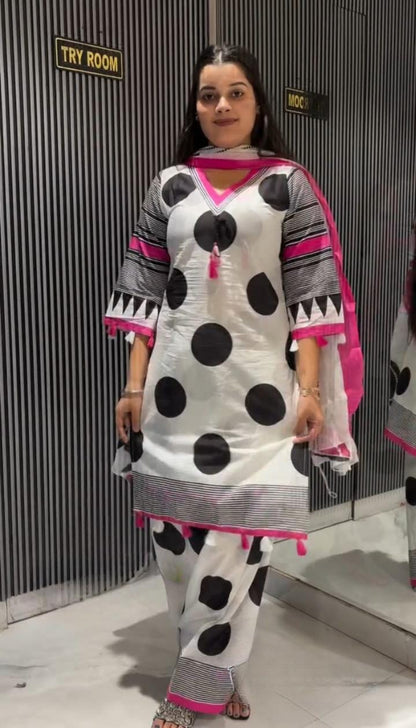 A woman modeling a white traditional outfit with large black polka dots, gray and black striped sleeve details, and bright pink accents, standing in front of a gray slatted wall.