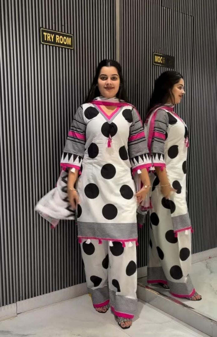 A woman modeling a white kurta set with black polka dots, gray patterned sleeves and hem, and bright pink trim, standing in front of a mirror that reflects her side profile. The background features a dark gray corrugated wall with a 'TRY ROOM' sign.
