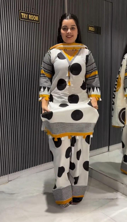 A woman modeling a white two-piece kurta set with black polka dots, gray stripes, and yellow accents, standing in front of a dark gray corrugated wall.