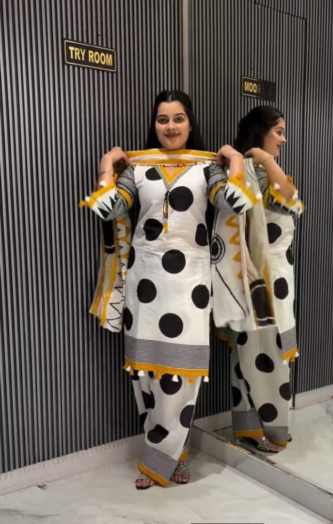 A woman modeling a white three-piece salwar kameez suit with black polka dots and yellow/gold accents, standing in front of a dark gray slatted wall with a mirror showing her reflection.