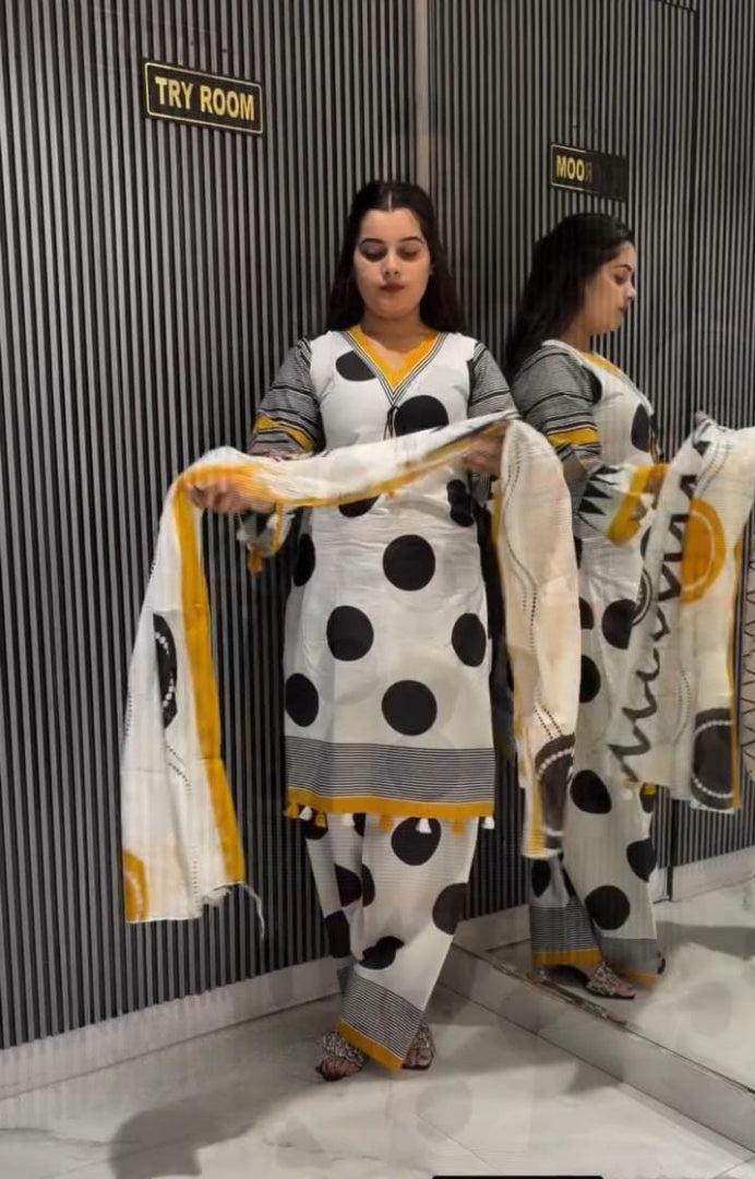 A woman stands in a fitting room wearing a white salwar suit with large black polka dots and gray sleeves, holding a matching dupatta with yellow and black accents. A mirror reflects her profile, and signs on the wall read 'TRY ROOM' and 'WOO'.